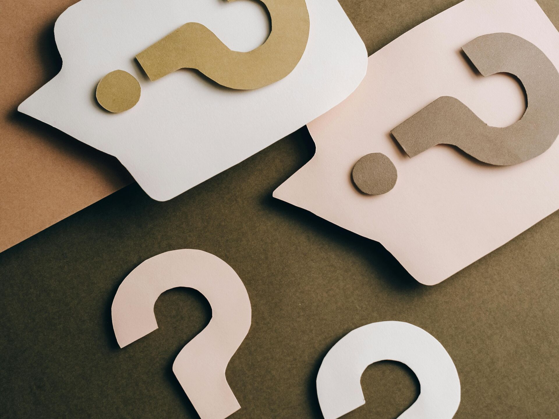 Papierausschnitte in Form von Sprechblasen und Fragezeichen in neutralen Farbtönen auf farbigem Untergrund.