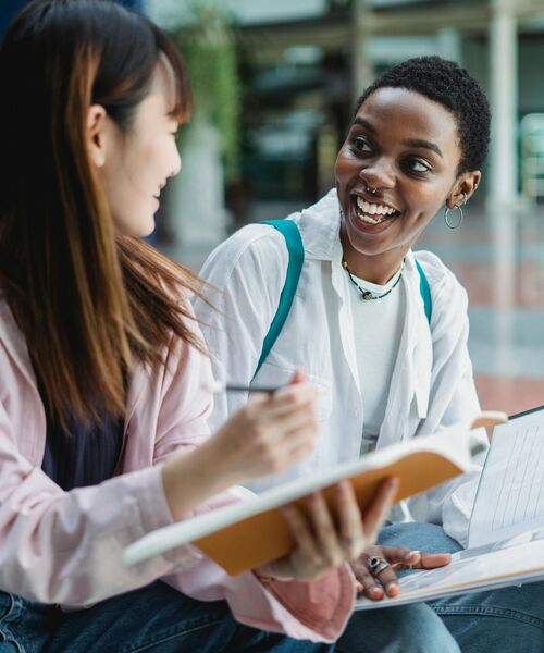 Zwei Studierende sitzen zusammen, unterhalten sich lachend und halten Notizbücher in den Händen.