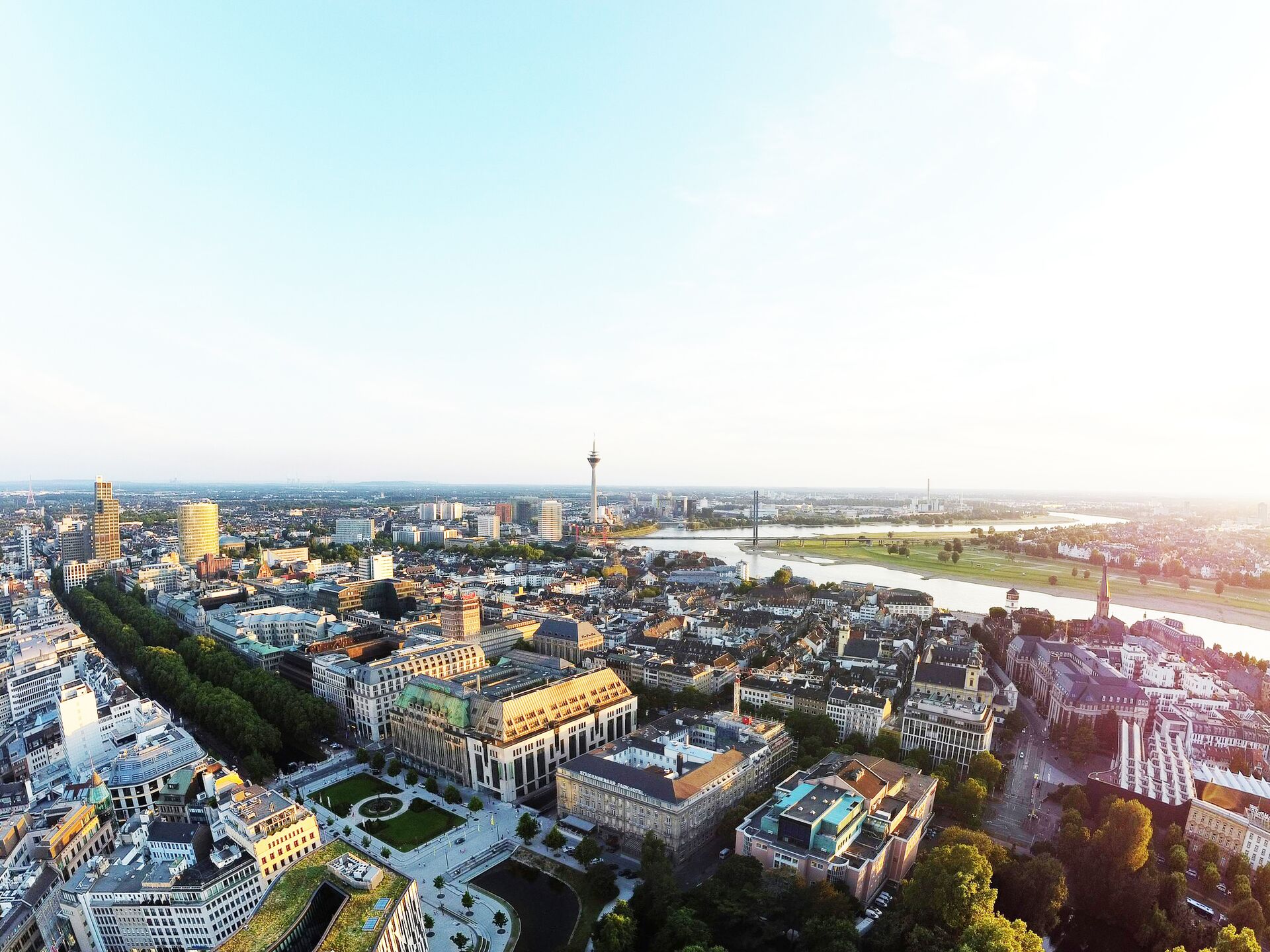 Luftaufnahme einer Stadt mit Fluss, Brücke und markantem Fernsehturm bei klarem Himmel.