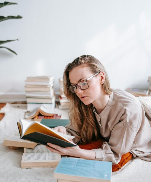 Person liegt entspannt auf dem Boden, liest ein Buch und ist umgeben von mehreren aufgeschlagenen Büchern.