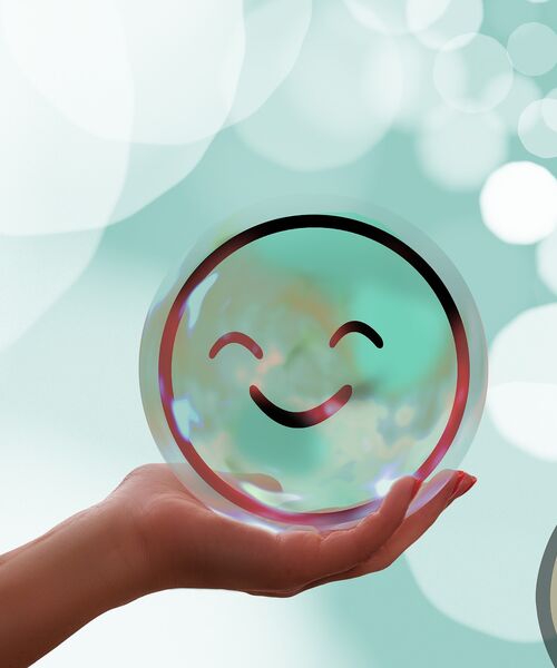 Hand hält eine schimmernde Kugel mit einem lächelnden Smiley auf hellem, weichgezeichnetem Hintergrund.