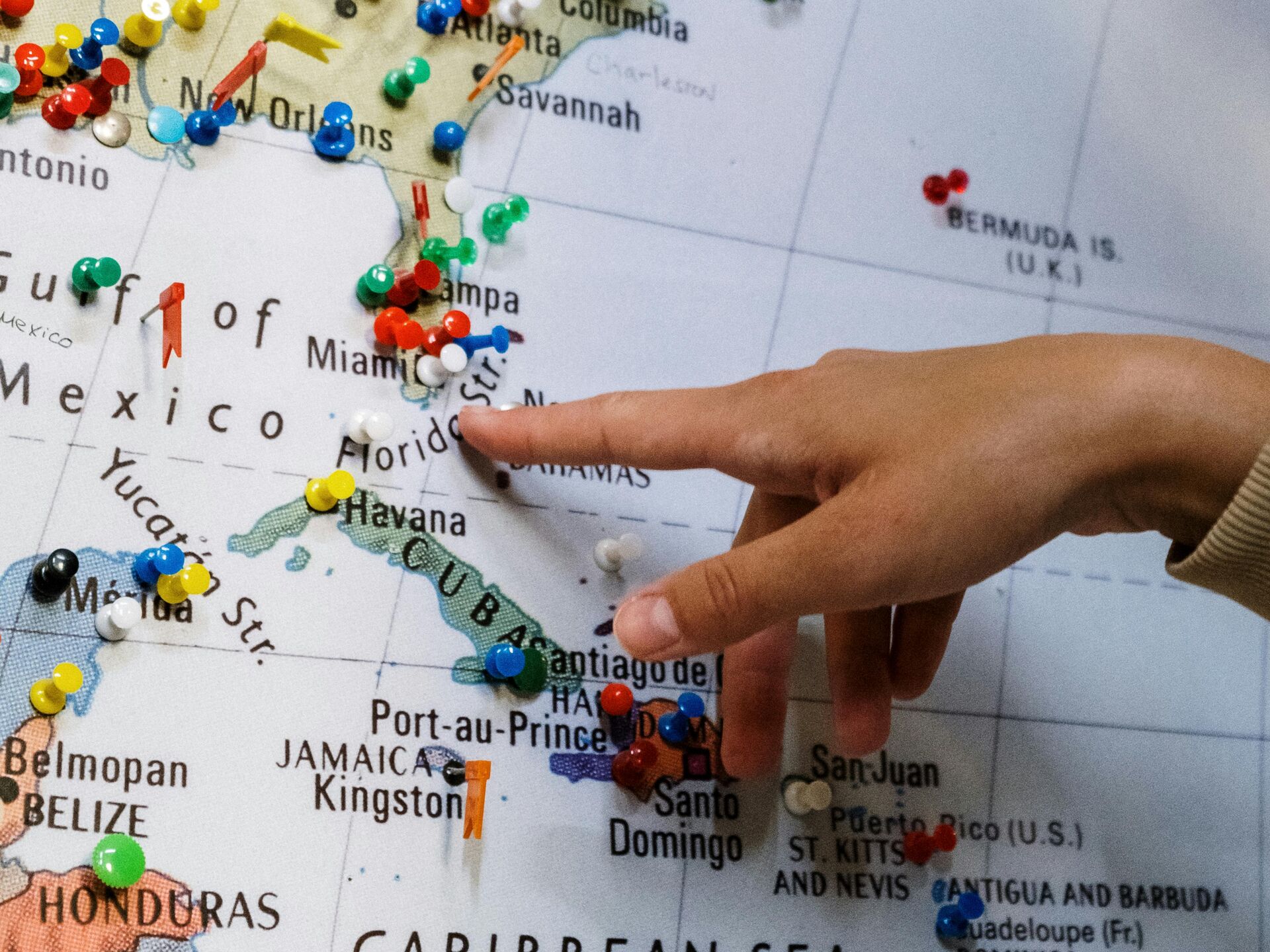 Hand zeigt auf eine Weltkarte mit vielen bunten Stecknadeln, die verschiedene Reiseziele markieren.