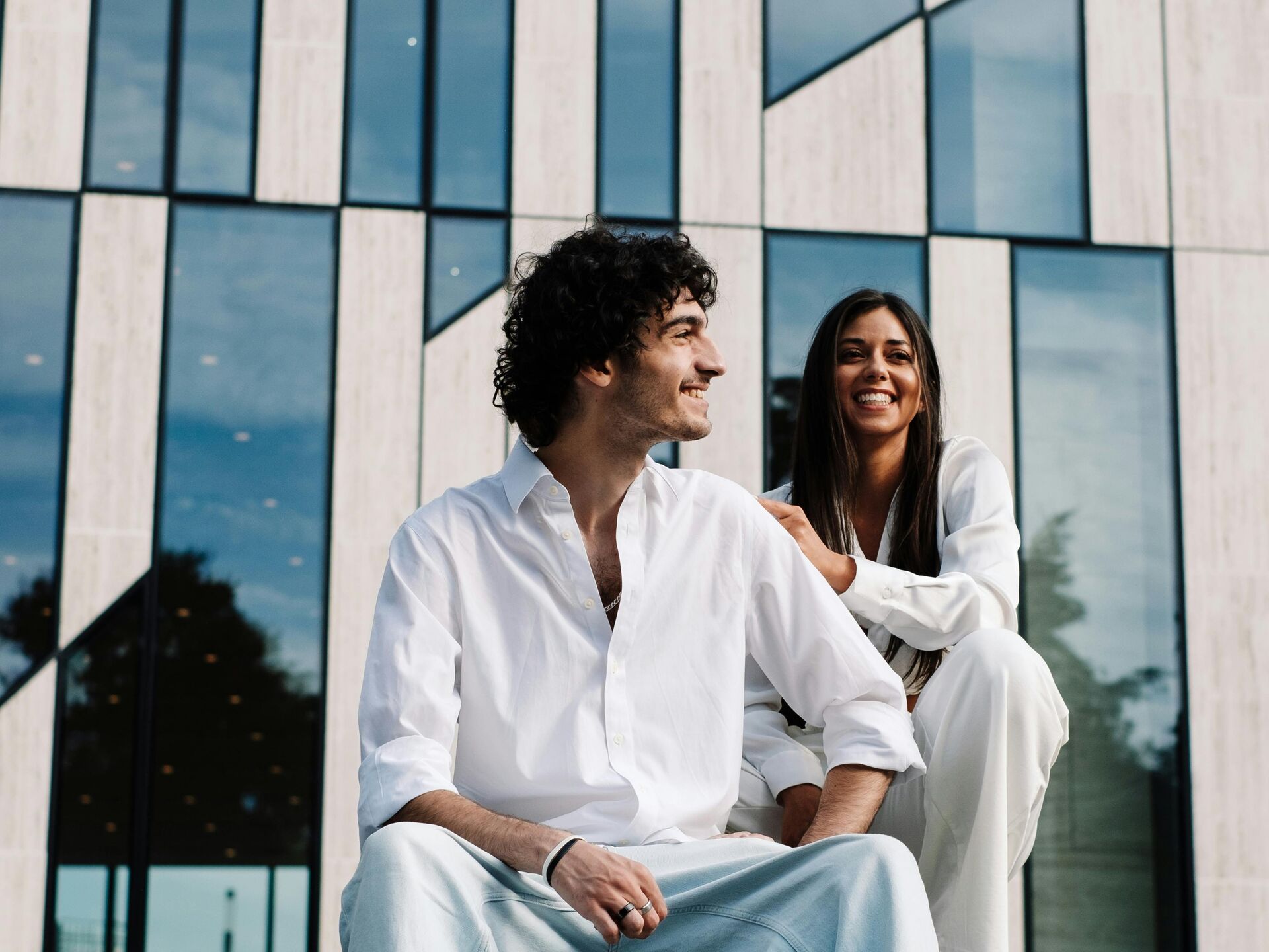 Zwei junge Menschen sitzen lachend auf einer Bank vor einem modernen Gebäude mit Glasfassade.