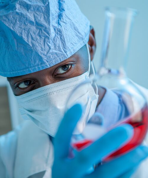 Forschungsperson in Schutzkleidung und Maske hält ein Laborgefäß mit roter Flüssigkeit.