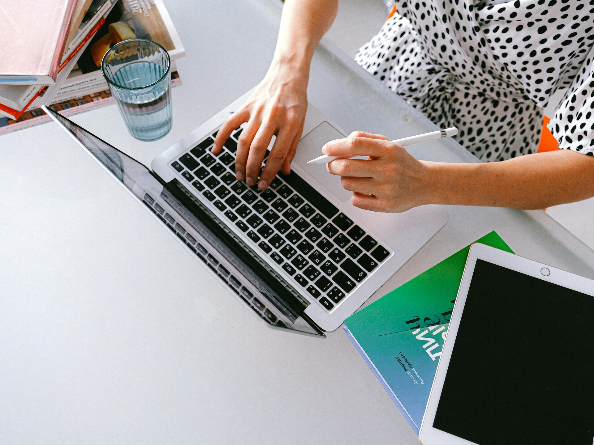 Person arbeitet an einem Laptop, hält einen Stift und hat Bücher sowie ein Tablet auf dem Tisch liegen.