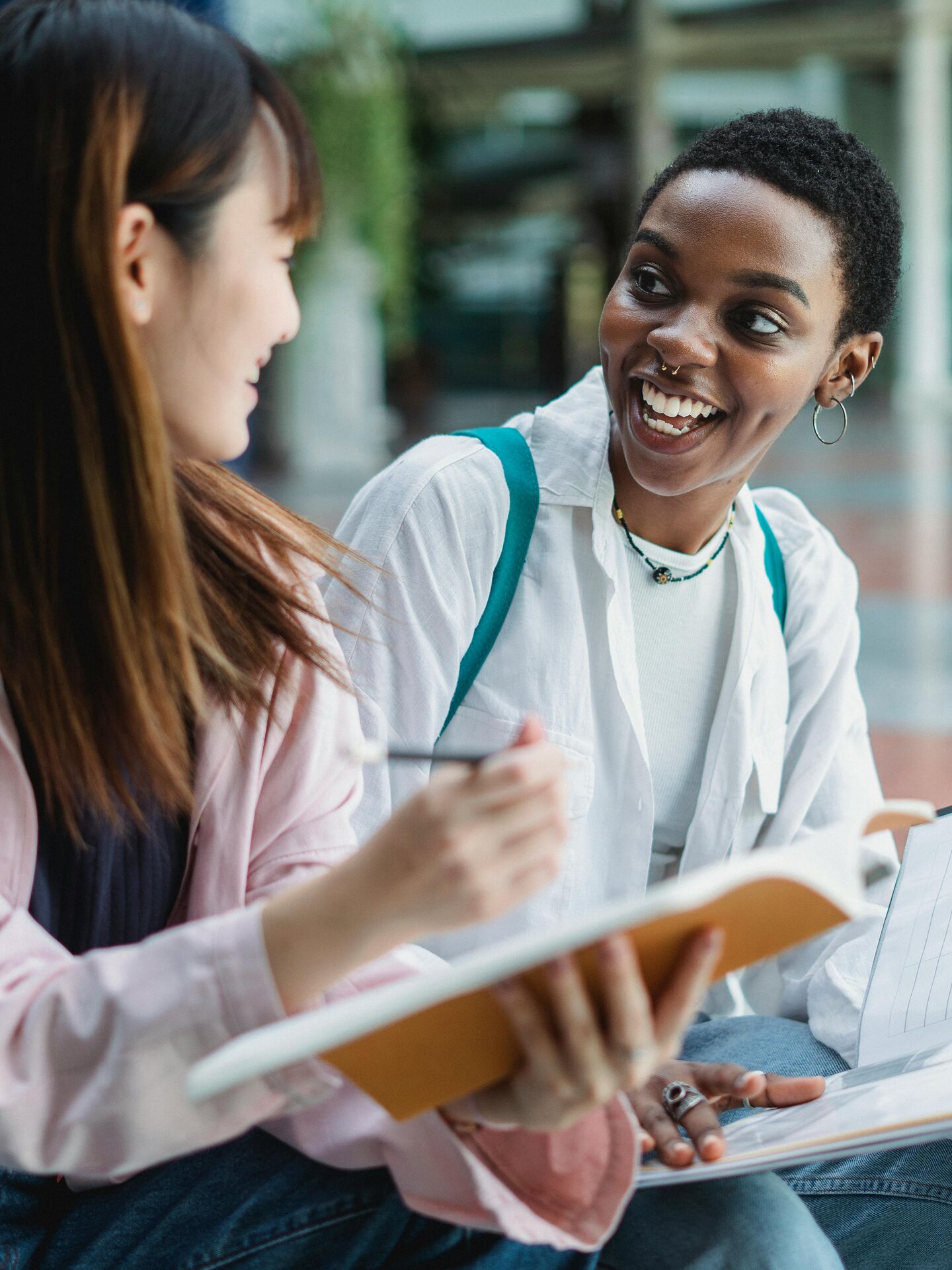 Zwei Studierende sitzen zusammen, unterhalten sich lachend und halten Notizbücher in den Händen.