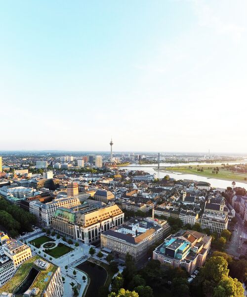 Luftaufnahme einer Stadt mit Fluss, Brücke und markantem Fernsehturm bei klarem Himmel.