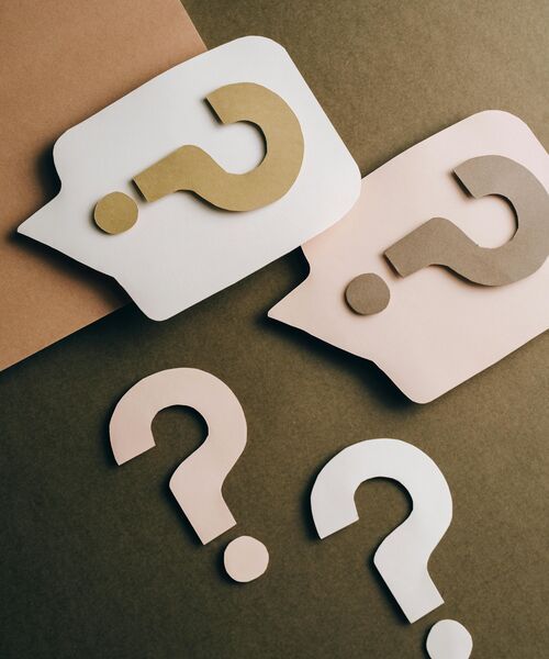 Papierausschnitte in Form von Sprechblasen und Fragezeichen in neutralen Farbtönen auf farbigem Untergrund.