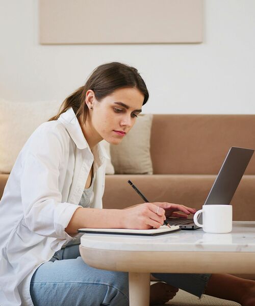 Person arbeitet konzentriert am Laptop und macht sich Notizen an einem Tisch mit Kaffeetasse.