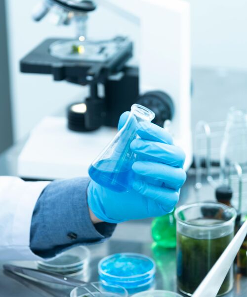 Laborfachkraft hält einen Kolben mit blauer Flüssigkeit, im Hintergrund steht ein Mikroskop.