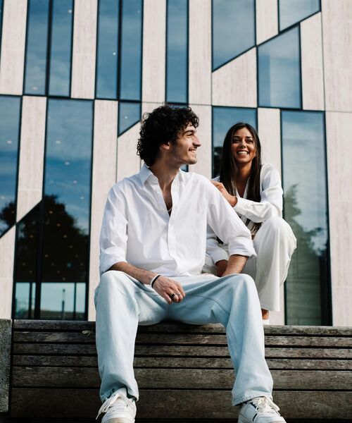 Zwei junge Menschen sitzen lachend auf einer Bank vor einem modernen Gebäude mit Glasfassade.