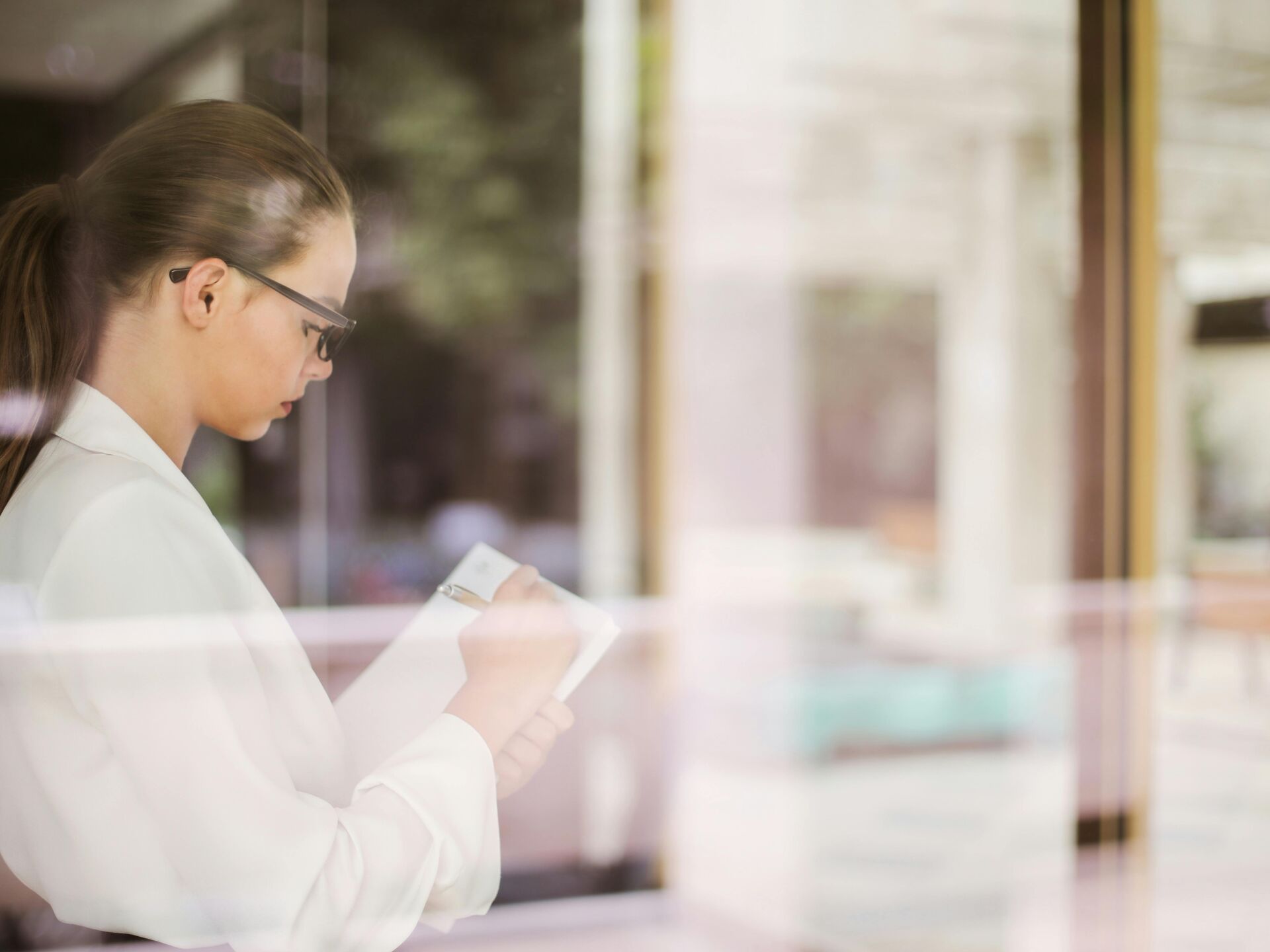 Frau schreibt konzentriert Notizen in ein Heft, im Hintergrund ein helles Büro mit Glasfront.