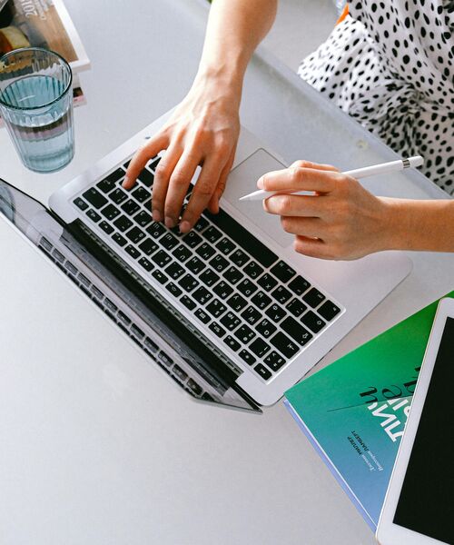 Person arbeitet an einem Laptop, hält einen Stift und hat Bücher sowie ein Tablet auf dem Tisch liegen.