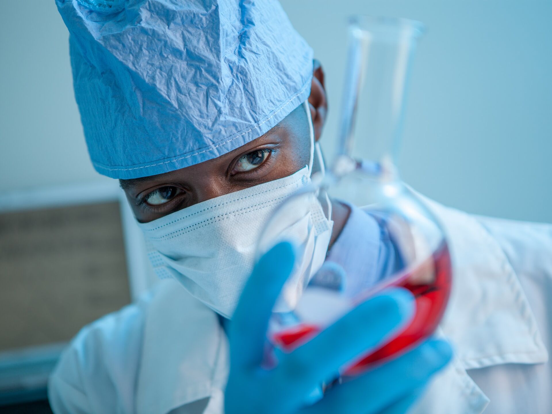 Forschungsperson in Schutzkleidung und Maske hält ein Laborgefäß mit roter Flüssigkeit.